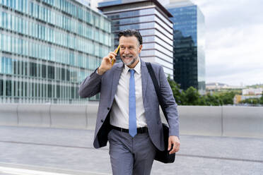 Smiling mature businessman talking through smart phone walking on street - OIPF02713