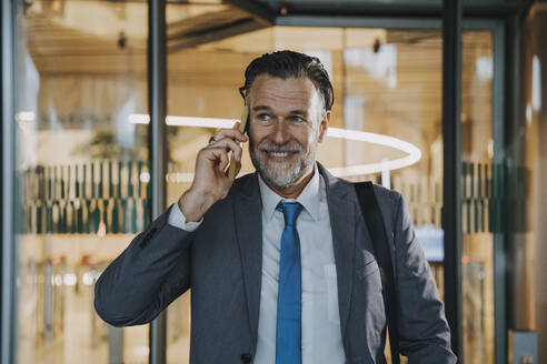 Lächelnder reifer Geschäftsmann, der vor einer Glastür mit einem Smartphone spricht - OIPF02719