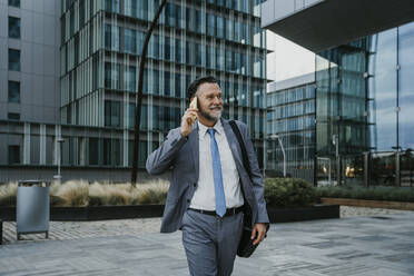 Smiling mature businessman talking through mobile phone walking on footpath - OIPF02720