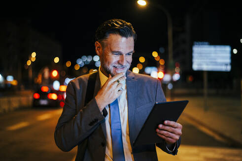 Nachdenklich lächelnder reifer Geschäftsmann stehend mit Tablet-PC auf der Straße - OIPF02737