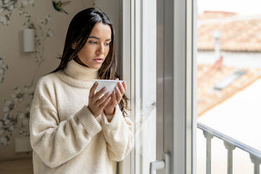 Nachdenkliche junge Frau steht mit einer Schüssel Suppe am Fenster ihres Hauses - DLTSF03488