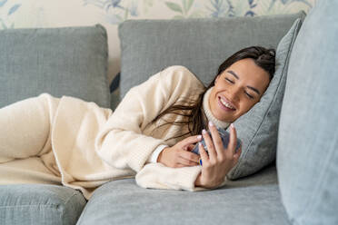 Happy young woman using smart phone on sofa at home - DLTSF03496