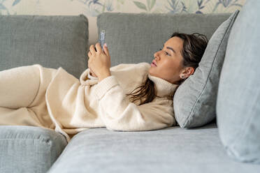 Junge Frau benutzt ihr Smartphone auf dem Sofa zu Hause - DLTSF03497