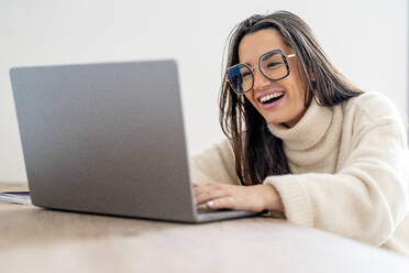 Glücklicher junger Freiberufler mit Laptop am Schreibtisch im Heimbüro - DLTSF03500