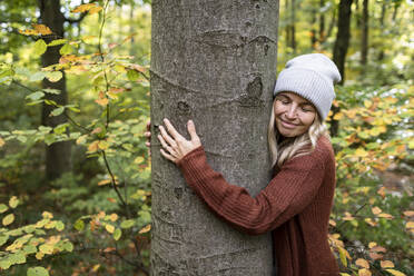 Smiling woman in knit hat embracing tree trunk at autumn forest - HMEF01437