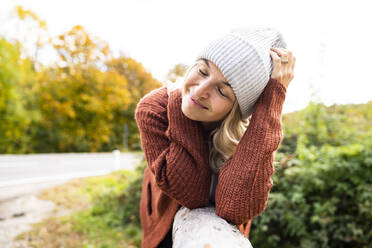 Smiling mature woman with eyes closed leaning on wood - HMEF01440