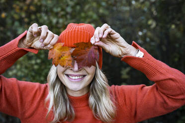 Smiling woman covering eyes with autumn leaves on weekend - HMEF01468