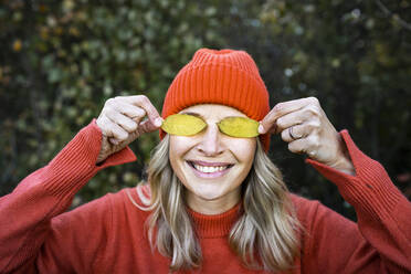 Smiling mature woman covering eyes with autumn leaves - HMEF01469