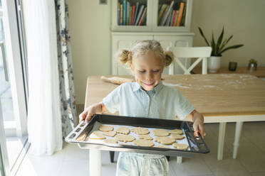 Smiling girl holding tray of cookies at home - SVKF00780