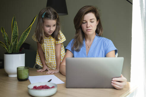 Geschäftsfrau arbeitet an einem Laptop, während ihre Tochter zu Hause am Schreibtisch steht - SVKF00797