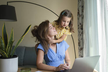 Cute daughter embracing mother sitting with laptop at desk in home office - SVKF00798