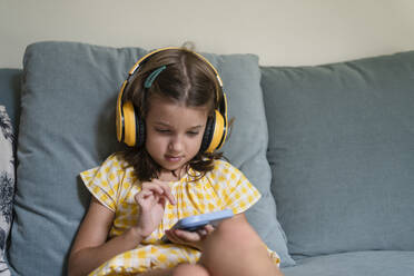 Girl wearing wireless headphones using smart phone on sofa at home - SVKF00805