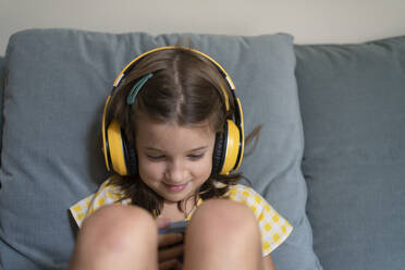 Smiling girl wearing wireless headphones using smart phone on sofa at home - SVKF00806