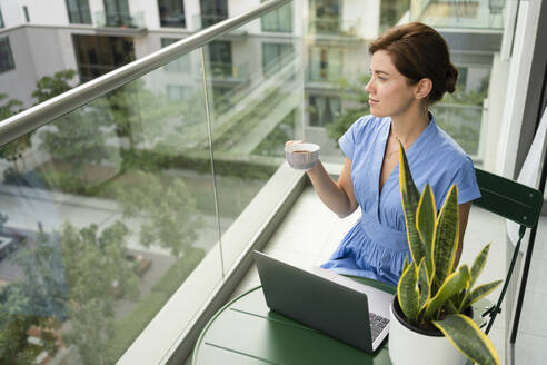 Businesswoman sitting with laptop and coffee cup in balcony - SVKF00813