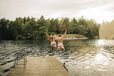 Male friends with arms raised jumping in lake from jetty during vacation - MASF32663