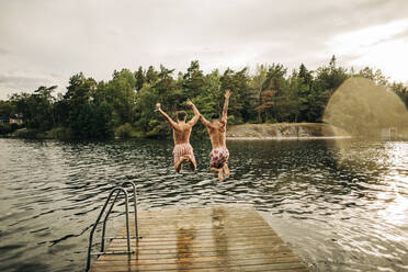 Carefree male friends with arms raised jumping in lake during vacation - MASF32665