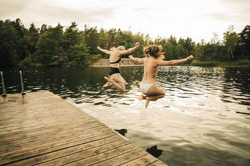 Female friends wearing bikini jumping in lake with arms outstretched during vacation - MASF32674