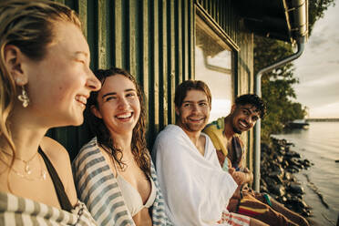 Happy male and female friends with towel sitting by cottage - MASF32691