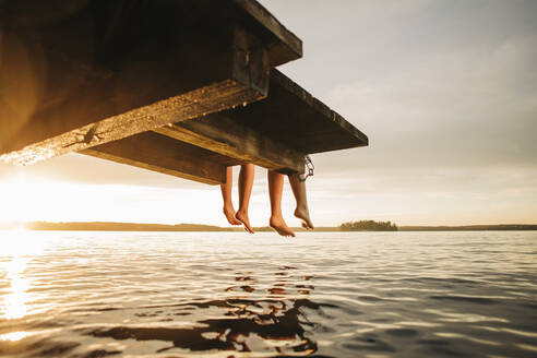 Niedriger Ausschnitt von Freundinnen, die bei Sonnenuntergang auf einem Steg am See sitzen - MASF32706