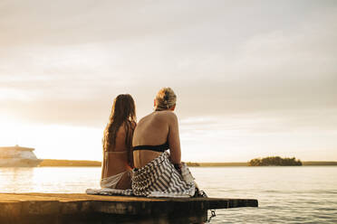 Rückansicht von Freundinnen, die bei Sonnenuntergang auf einem Steg sitzen und auf den See blicken - MASF32707
