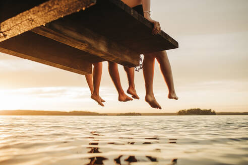 Niedriger Ausschnitt von männlichen und weiblichen Freunden, die bei Sonnenuntergang auf einem Steg am See sitzen - MASF32709