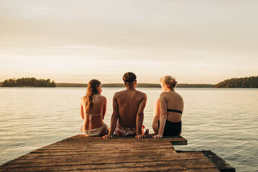 Rückansicht von männlichen und weiblichen Freunden, die im Urlaub auf einem Steg am See sitzen - MASF32710