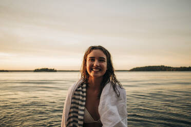 Porträt einer glücklichen Frau mit Handtuch am See bei Sonnenuntergang - MASF32716