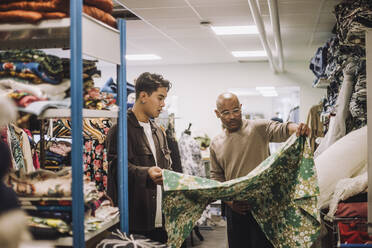 Männlicher Modedesigner mit einem Kollegen, der in einer Werkstatt Stoffe untersucht - MASF32757