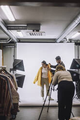 Fotograf fotografiert Designer hilft Modell tragen Jacke vor weißem Hintergrund im Studio - MASF32760