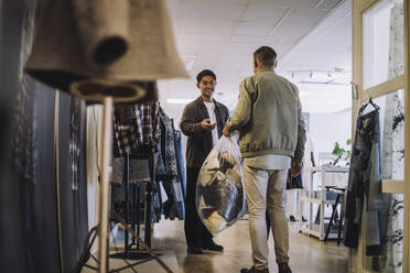 Rückansicht eines männlichen Modedesigners, der einem Kollegen in einem Workshop eine Plastiktüte mit recycelter Kleidung übergibt - MASF32804