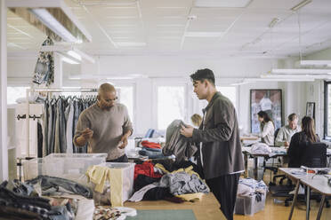 Male fashion designers discussing with each other while sorting recycled clothes at workshop - MASF32815