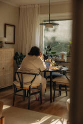 Rückansicht einer jungen Frau, die mit einem Laptop auf einem Stuhl zu Hause sitzt - MASF32842
