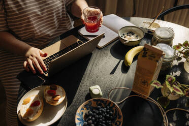 High angle view of woman holding drinking glass while using laptop with breakfast on table at home - MASF32877