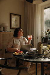 Junge Frau hält ein Trinkglas in der Hand und sitzt mit einem Laptop am Tisch - MASF32888