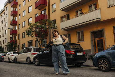 Porträt einer jungen selbstbewussten Frau, die mit der Hand auf der Hüfte vor einem Auto steht - MASF32893