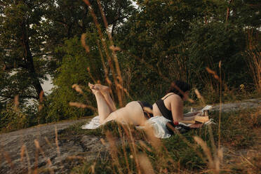 Full length of young woman in bikini reading book while lying against trees - MASF32918