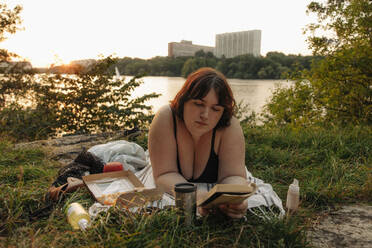 Junge Frau liest ein Buch, während sie bei Sonnenuntergang am See liegt - MASF32919