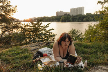 Young voluptuous woman reading book while lying against lake - MASF32920