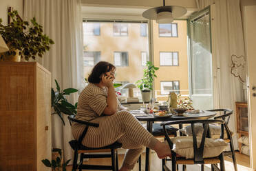 Young woman talking on smart phone while sitting on chair at home - MASF32921