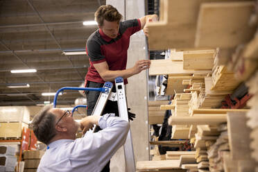 Verkäufer steht auf einer Leiter und entfernt ein Brett für einen männlichen Kunden im Baumarkt - MASF32935