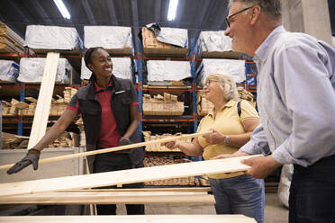 Smiling saleswoman looking at male and female customer holding planks at hardware store - MASF32946