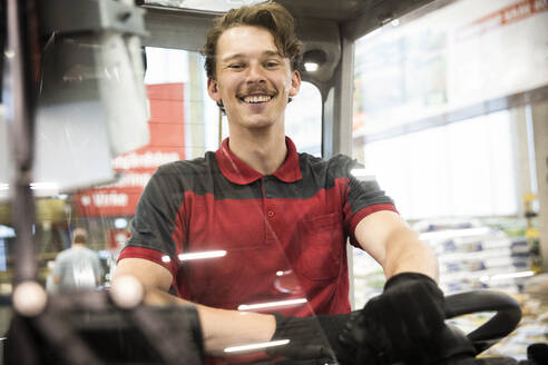 Portrait of happy male warehouse worker sitting in forklift seen through windshield - MASF32989