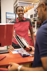 Smiling female cashier looking at male customer holding merchandise at checkout counter - MASF33016