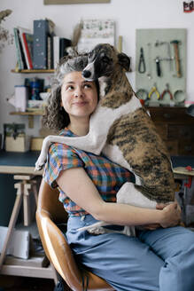 Lächelnde Frau, die ihren Hund umarmt, während sie zu Hause auf einem Stuhl sitzt - MASF33024
