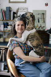 Porträt einer lächelnden Frau, die ihren Hund umarmt, während sie zu Hause auf einem Stuhl sitzt - MASF33025