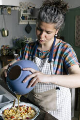 Frau gießt Teig aus einer Schüssel auf zerkleinerte Früchte in der Küche - MASF33039