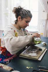 Weibliche Kunsthandwerkerin hält einen Krug, während sie zu Hause Schmuck herstellt - MASF33057
