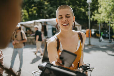 Smiling woman with shaved head paying via tap to pay while shopping at flea market - MASF33133