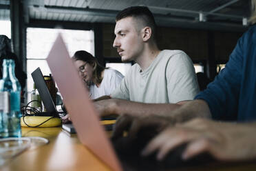 Konzentrierter Geschäftsmann arbeitet am Laptop inmitten von Kollegen in einem Start-up-Unternehmen - MASF33187