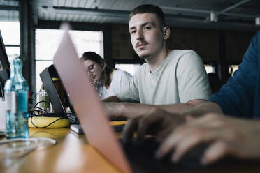 Porträt eines Geschäftsmannes mit Laptop, der inmitten von Kollegen im Büro sitzt - MASF33188